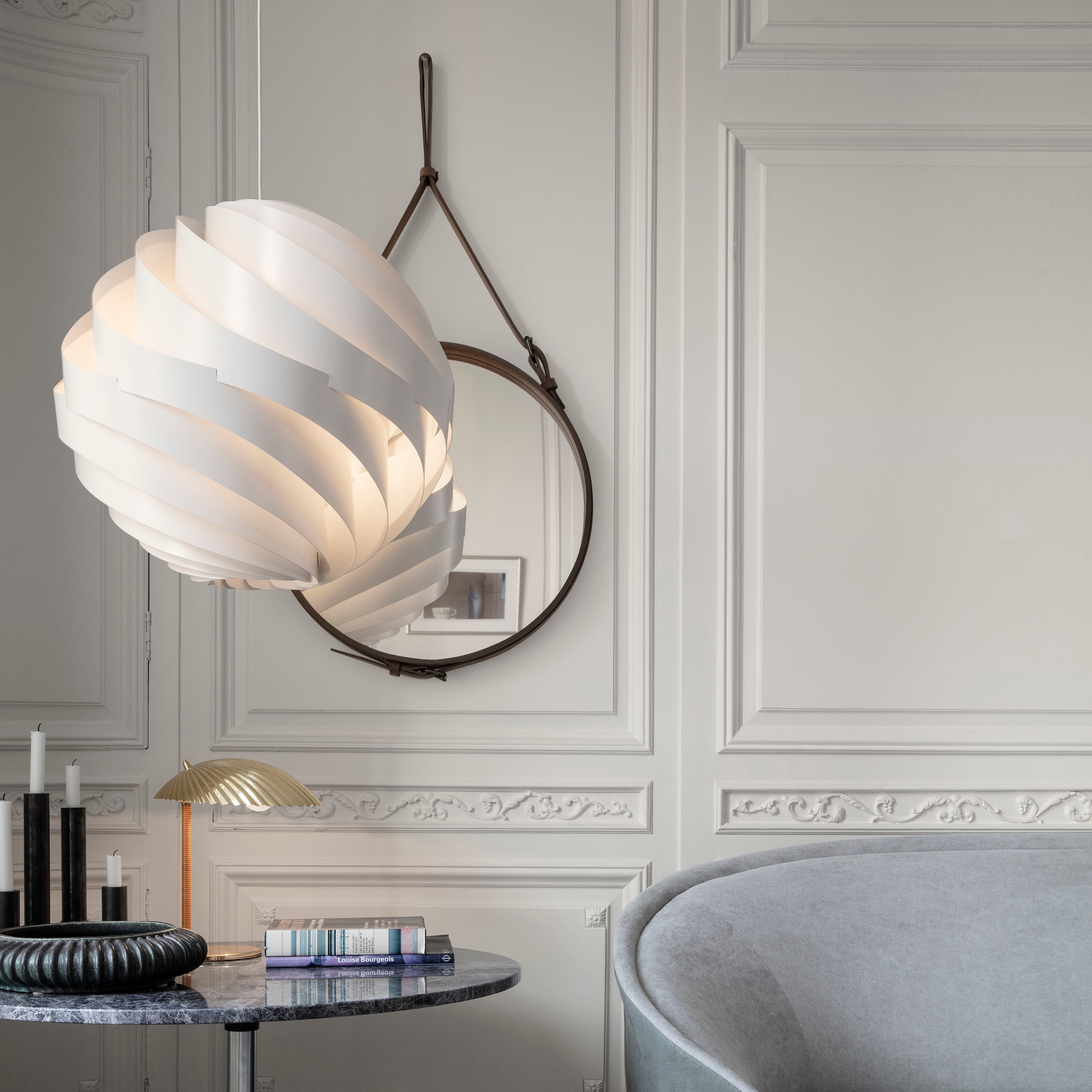 A round mirror with a brown leather strap hangs on a panelled wall beside a white spiral pendant lamp