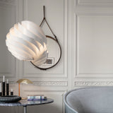 A round mirror with a brown leather strap hangs on a panelled wall beside a white spiral pendant lamp