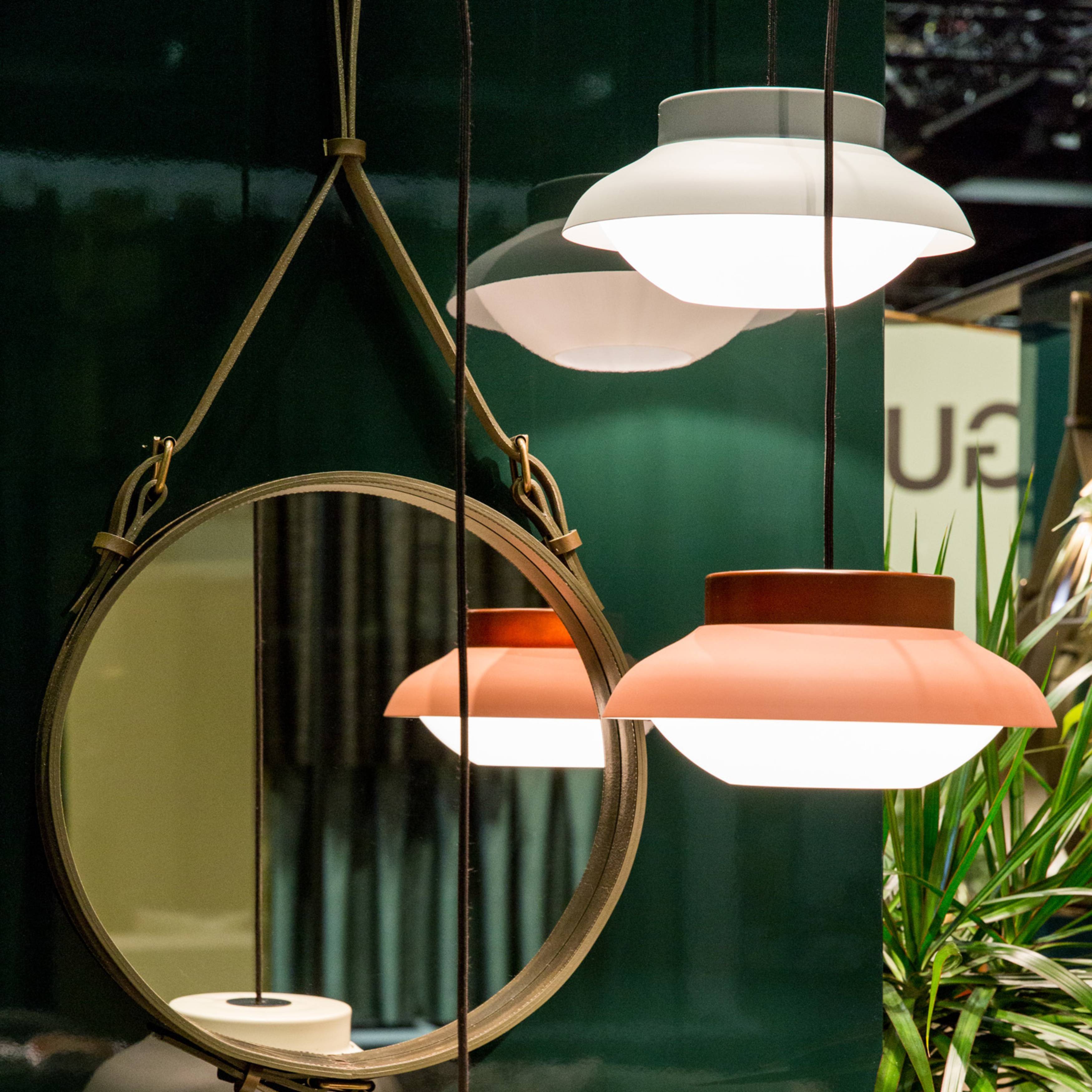 A round mirror hung from olive leather straps beside glowing pink and white pendant lamps on a green wall