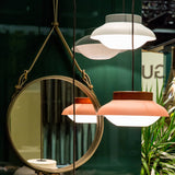 A round mirror hung from olive leather straps beside glowing pink and white pendant lamps on a green wall