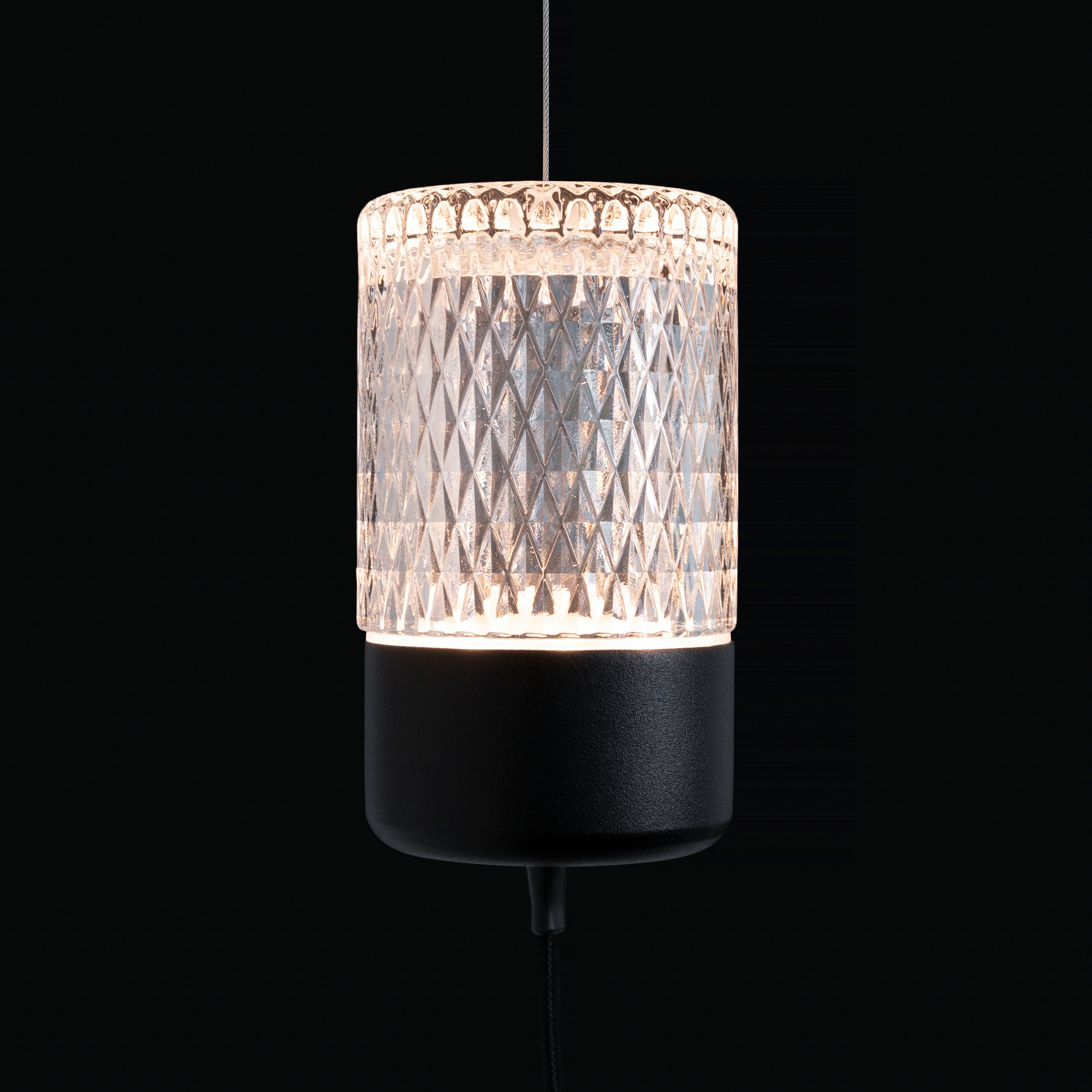 A glowing cylindrical pendant of clear diamond-patterned glass on a black base, hung from a thin cable