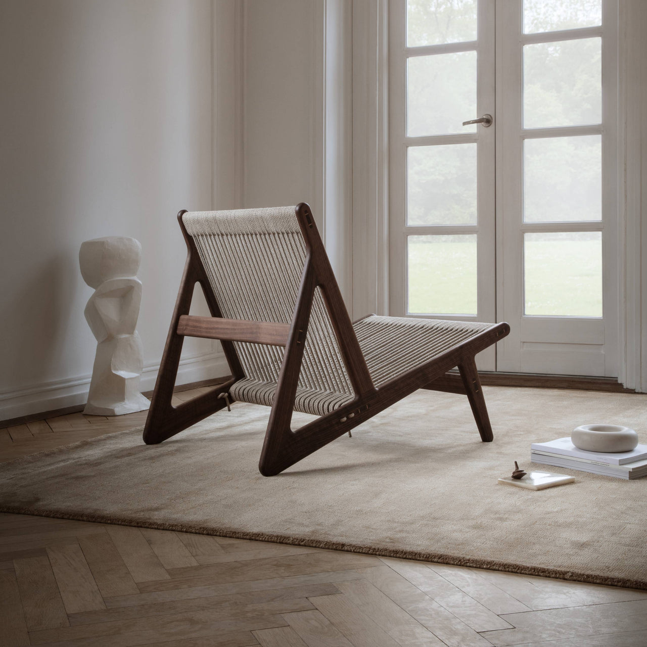 A low lounge chair with a dark wood triangular frame and woven rope seat on a beige rug