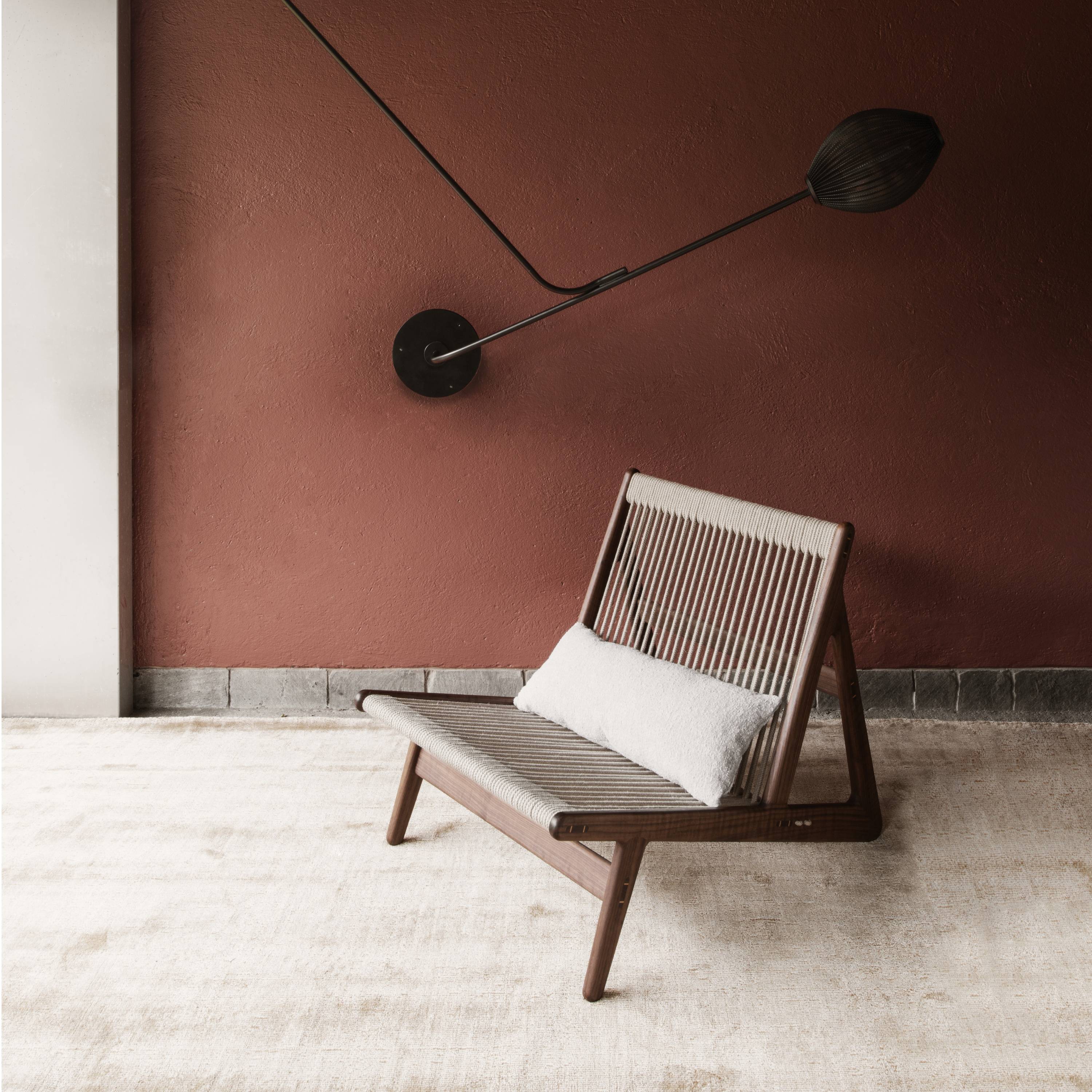 A low wooden lounge chair with cream cord seat and back and a white cushion, before a rust wall