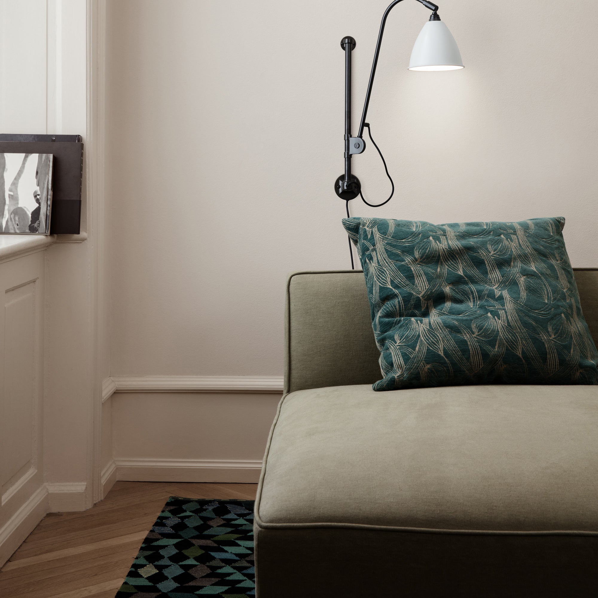 A low daybed in sage green fabric with a teal patterned cushion, beside a wall lamp and a geometric rug