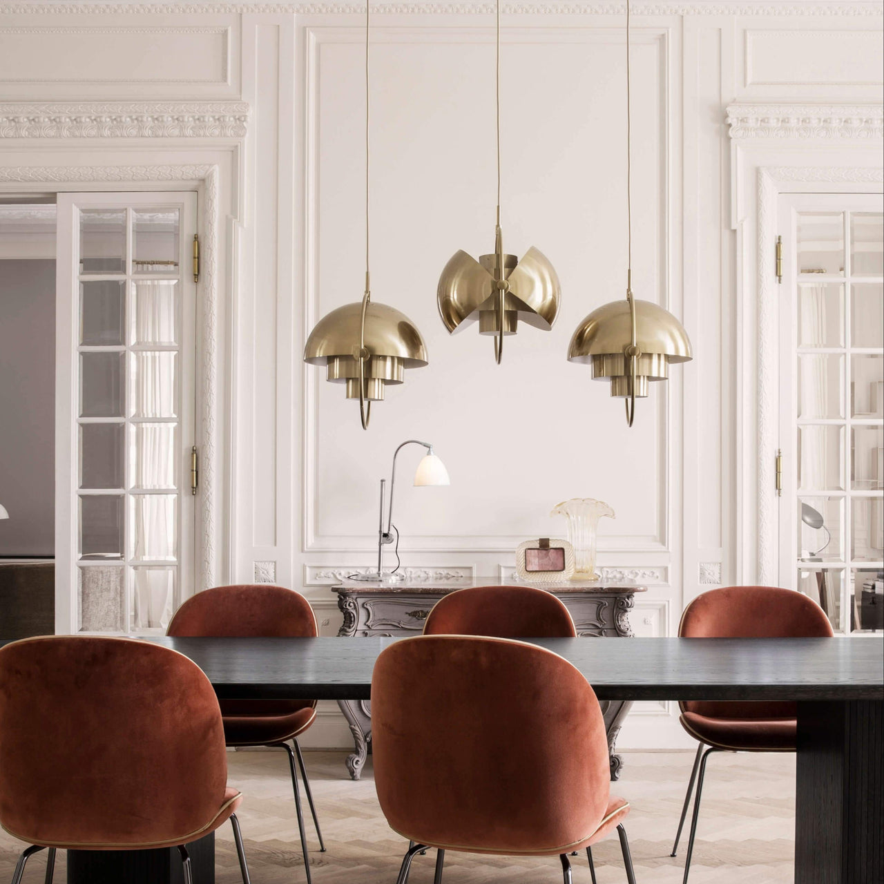 Three brass dome pendants with tiered inner shades hang over a dark table and rust velvet chairs