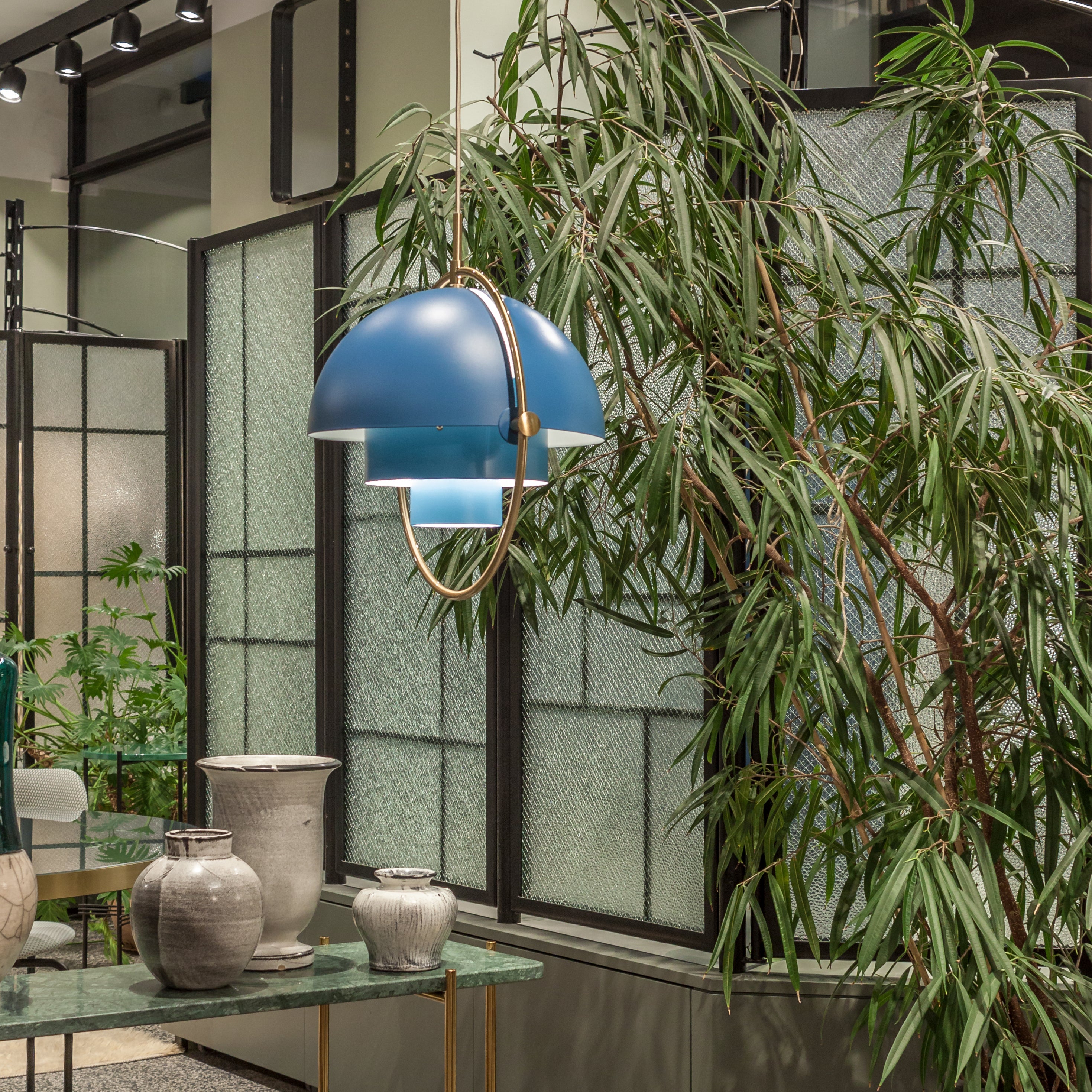 A blue tiered pendant with a brass ring hangs beside a leafy plant and textured glass panels