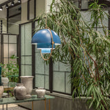 A blue tiered pendant with a brass ring hangs beside a leafy plant and textured glass panels