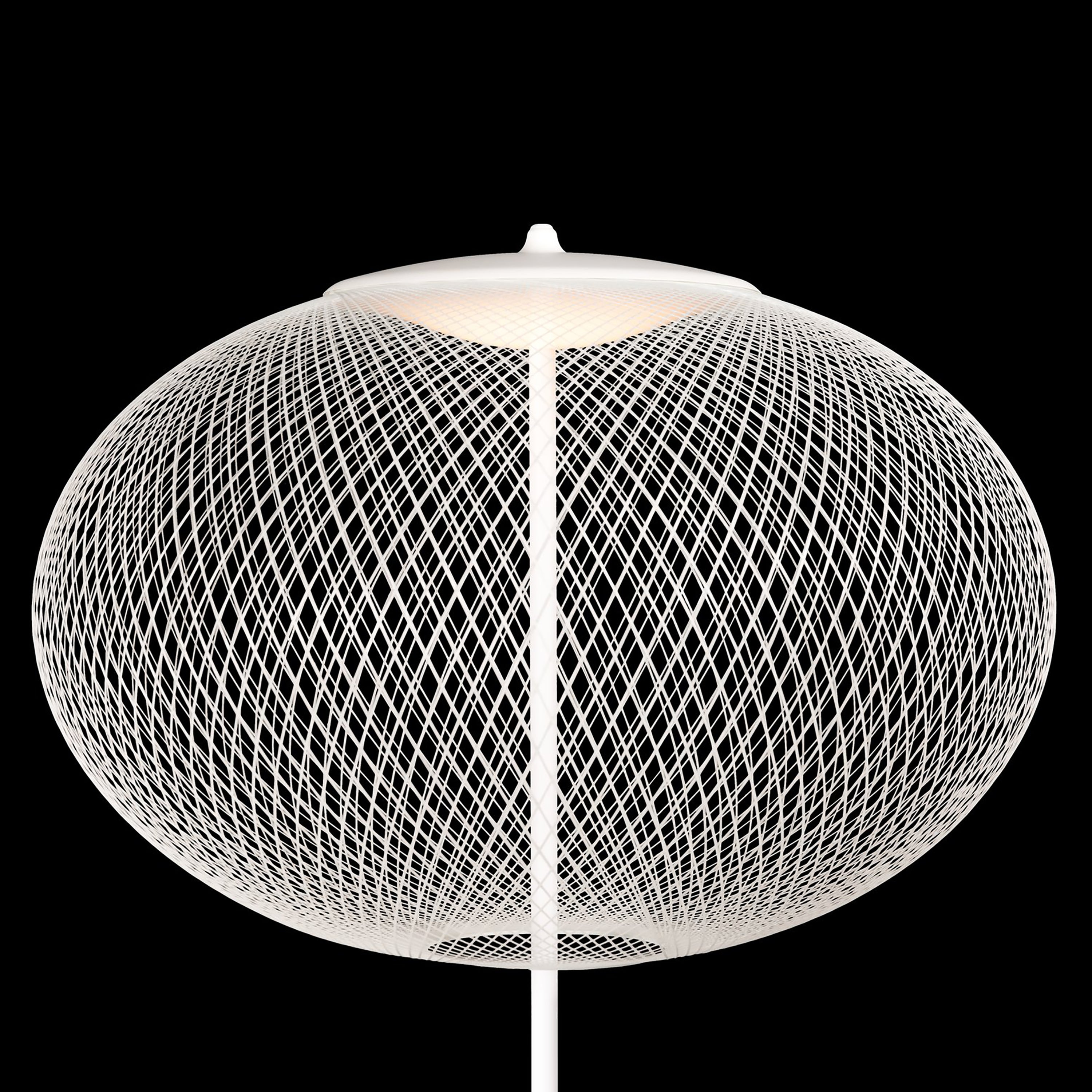 A white floor lamp with a wide oval shade of fine crisscrossing wire, lit from above, on black