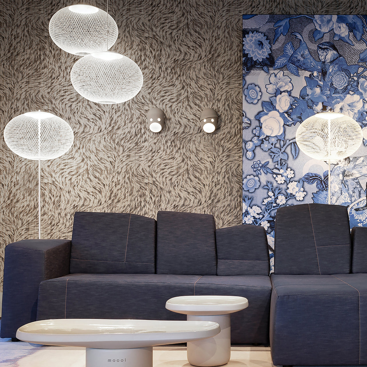 A white wire-mesh floor lamp with an oval shade beside pendants, in a room with a dark blue sofa