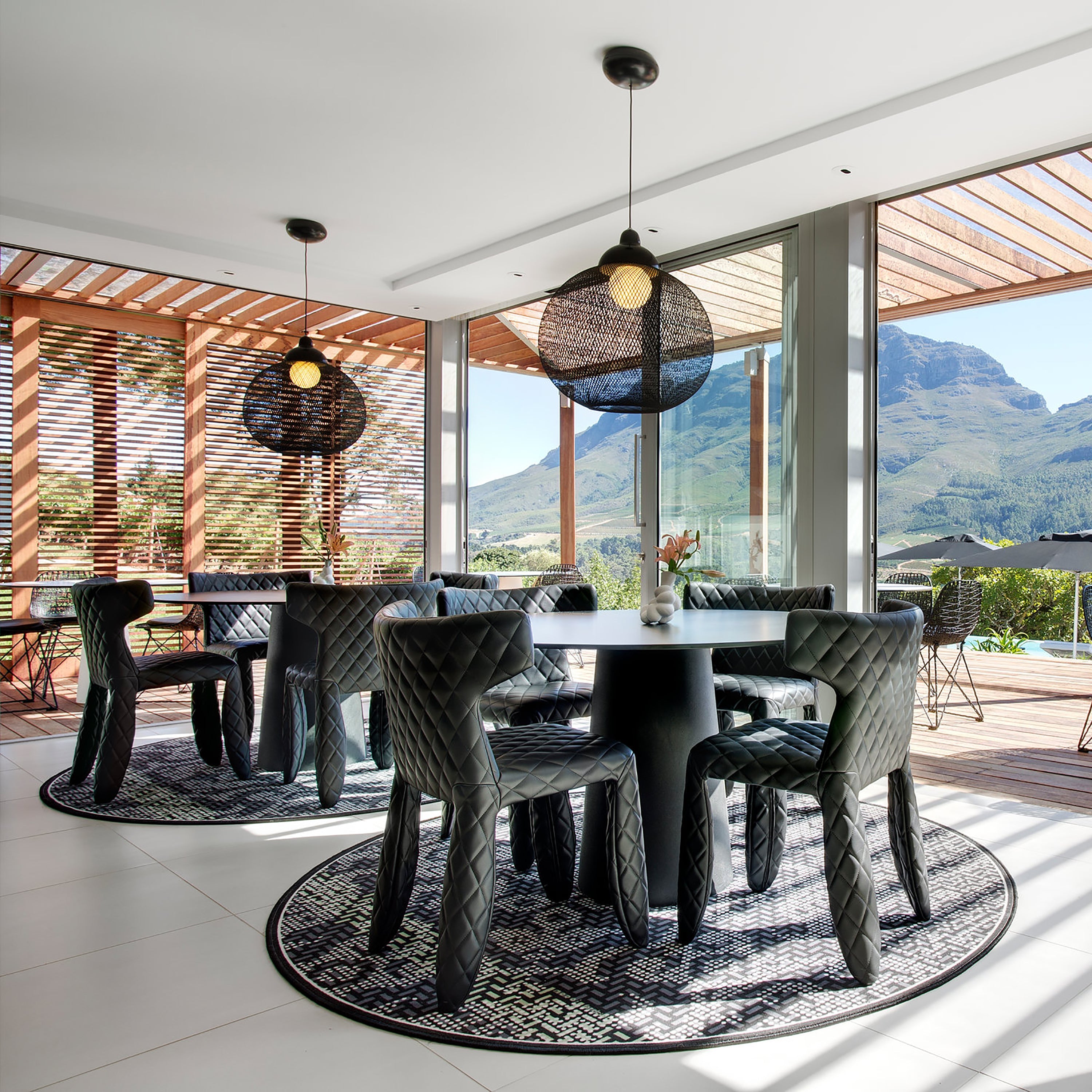 Round black woven pendant lamps above dark quilted chairs at round tables, with mountain views