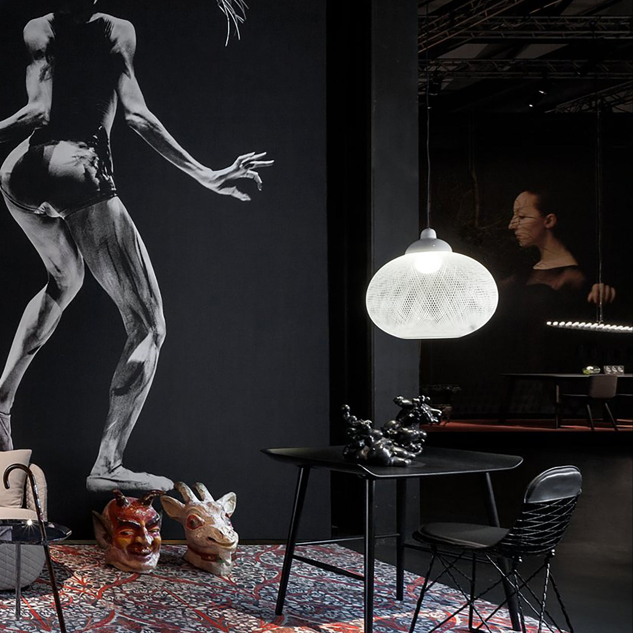 A round white mesh pendant lamp glows above a black table and wire chair, beside a large black-and-white mural