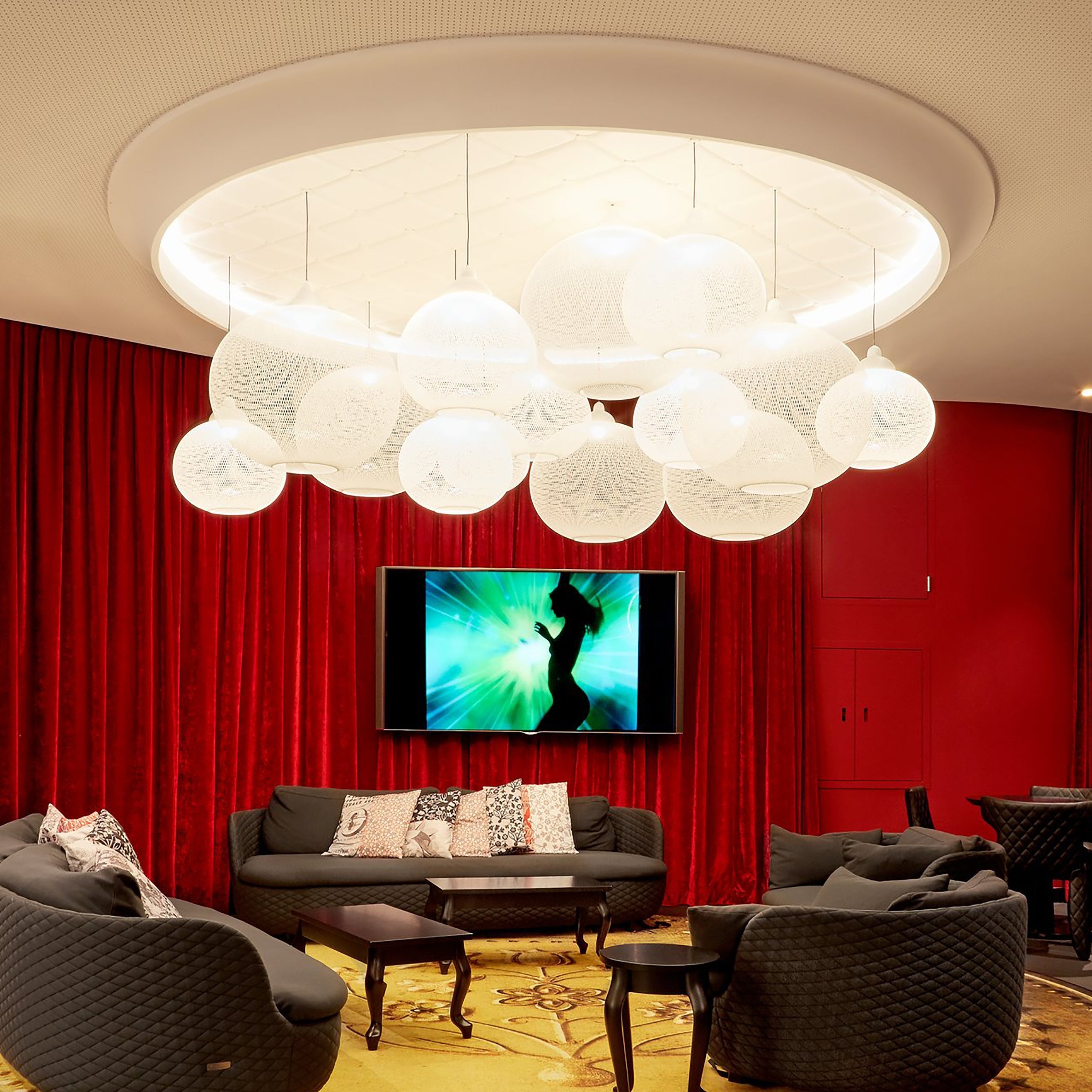 A cluster of white woven globe pendants hangs in a round ceiling recess above grey sofas and red curtains