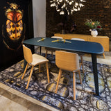 Light wood dining chairs around a dark teal table on a patterned rug, beside a brick wall