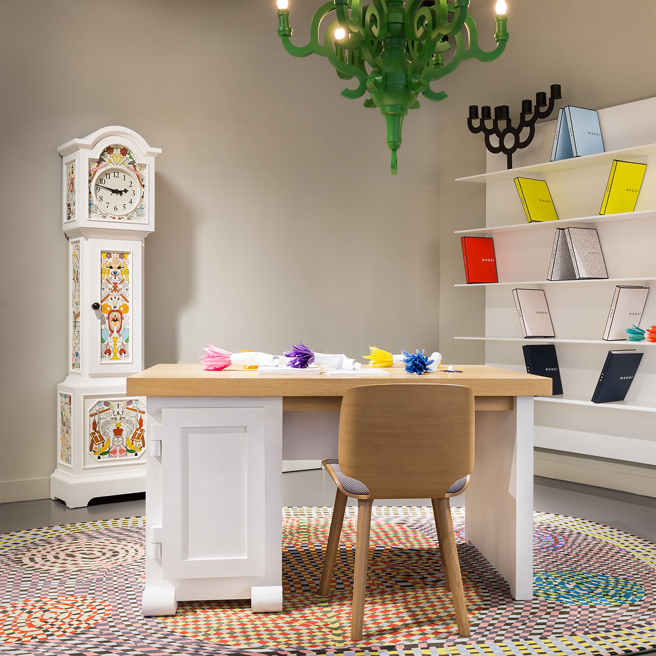 A light oak dining chair at a white desk with a patterned tall clock and green chandelier